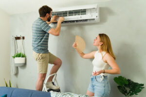 Woman fanning herself as her partner checks broken indoor AC unit.
