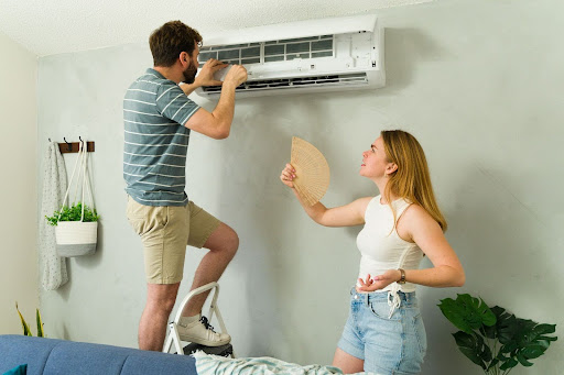 Woman fanning herself as her partner checks broken indoor AC unit.
